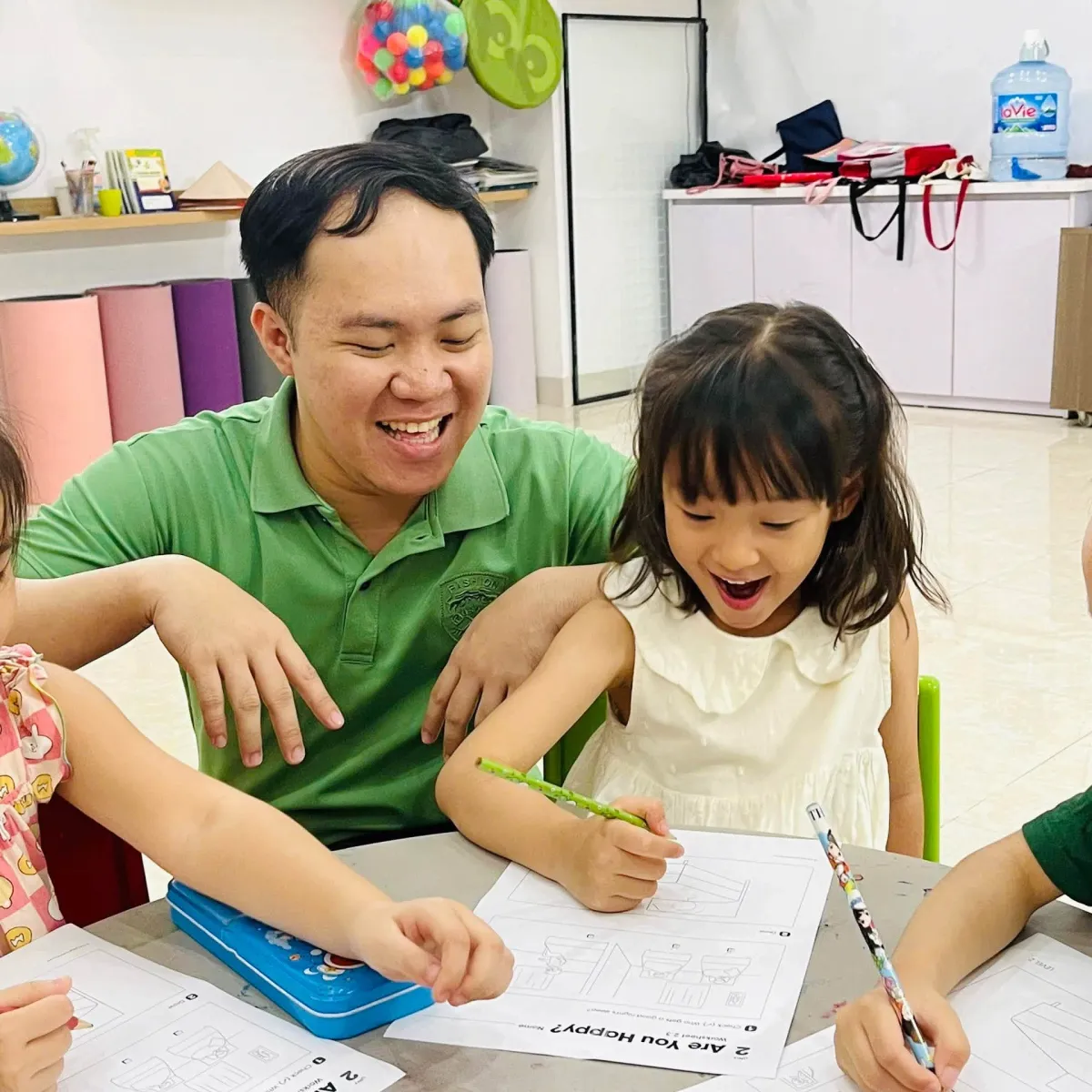 Thầy giáo cười vui cùng một bạn nhỏ đang làm phiếu bài tập trong lớp học tại Clever Junior Long Biên