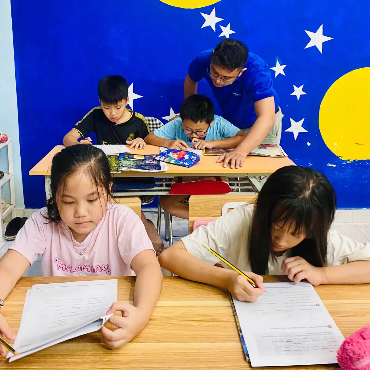 Thầy giáo cúi xuống hướng dẫn các bạn nhỏ làm bài trong lớp học tại Clever Junior Long Biên