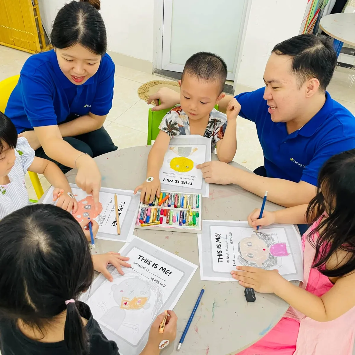 Hai thầy cô cùng các bạn nhỏ ngồi quanh bàn tô màu phiếu bài tập This is me!