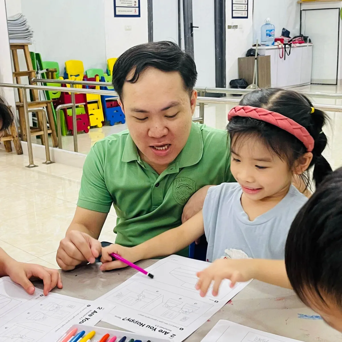 Thầy giáo ngồi cạnh bạn nhỏ làm phiếu bài tập Are You Happy?