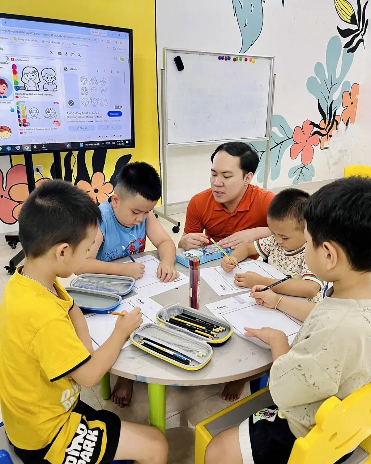 Thầy giáo cùng nhóm các bạn nam ngồi quanh bàn tròn làm phiếu bài tập Faces, màn hình phía sau chiếu hình gương mặt