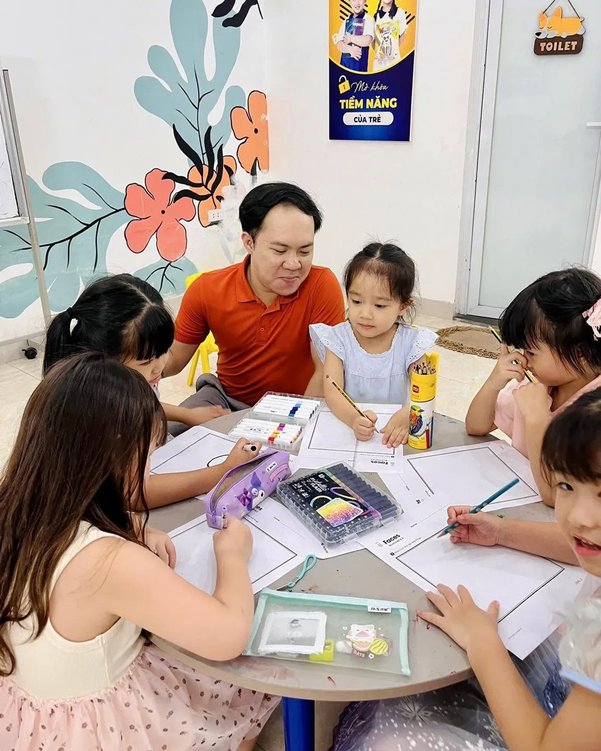 Thầy giáo ngồi cạnh một bạn nhỏ đang tô phiếu bài tập ở bàn tròn cùng các bạn tại Clever Junior Long Biên