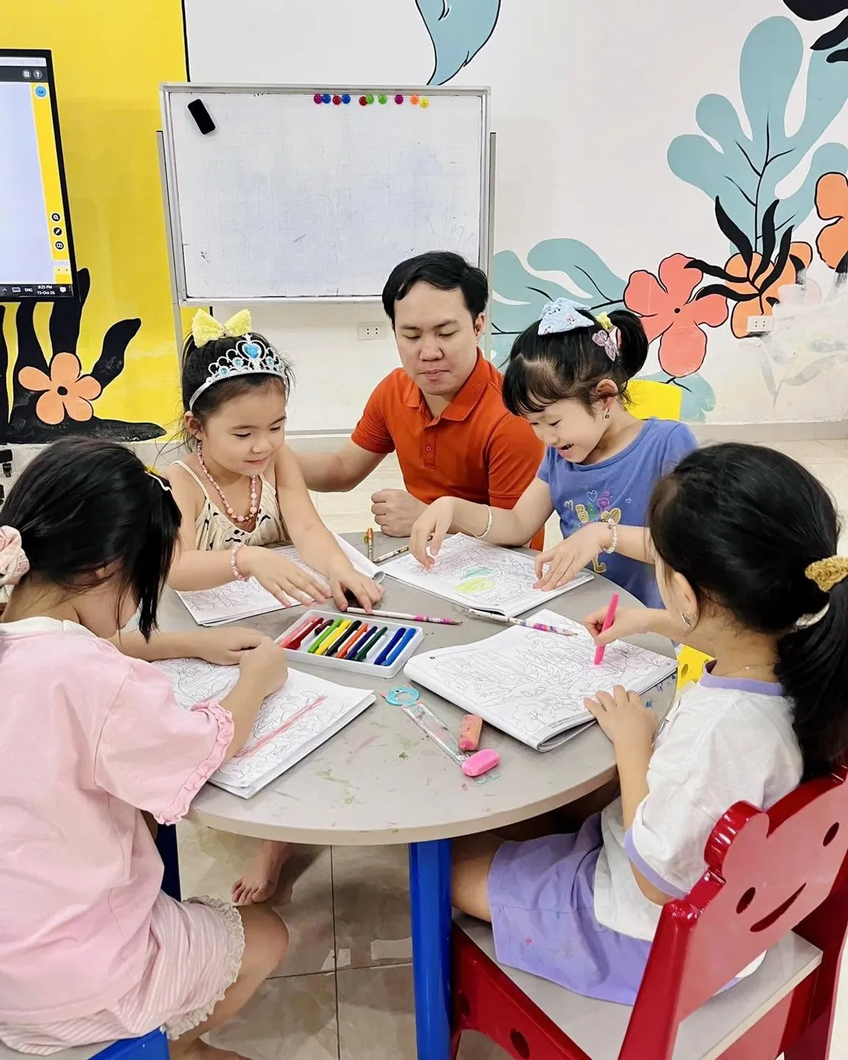 Các bạn nhỏ tô màu sách tranh quanh bàn tròn, thầy giáo ngồi cùng bàn tại Clever Junior Long Biên